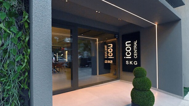 Egy modern épület, üveg homlokzattal, rajta az „icon SKG CENTRAL” felirattal, és az előtte lévő, gondosan nyírt bokorral.