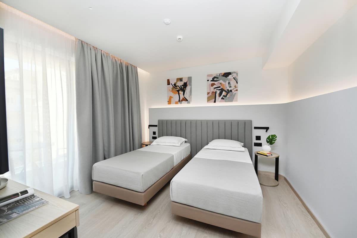 A modern hotel room with two single beds, a desk, a lamp, and abstract wall art.