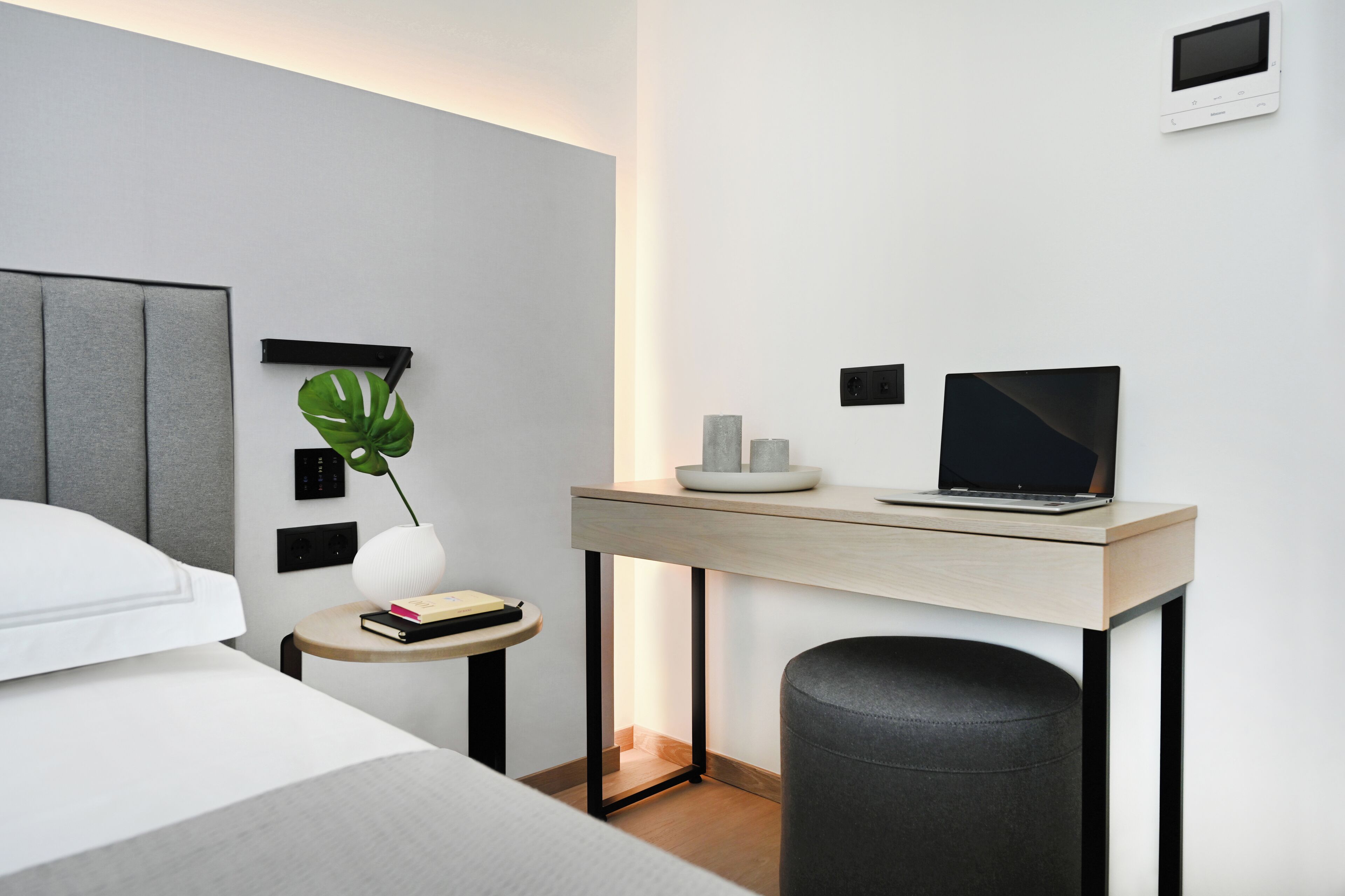 Una habitación de hotel moderna con una cama, un escritorio con una computadora portátil, una mesita con un libro, una planta y un cesto de basura.