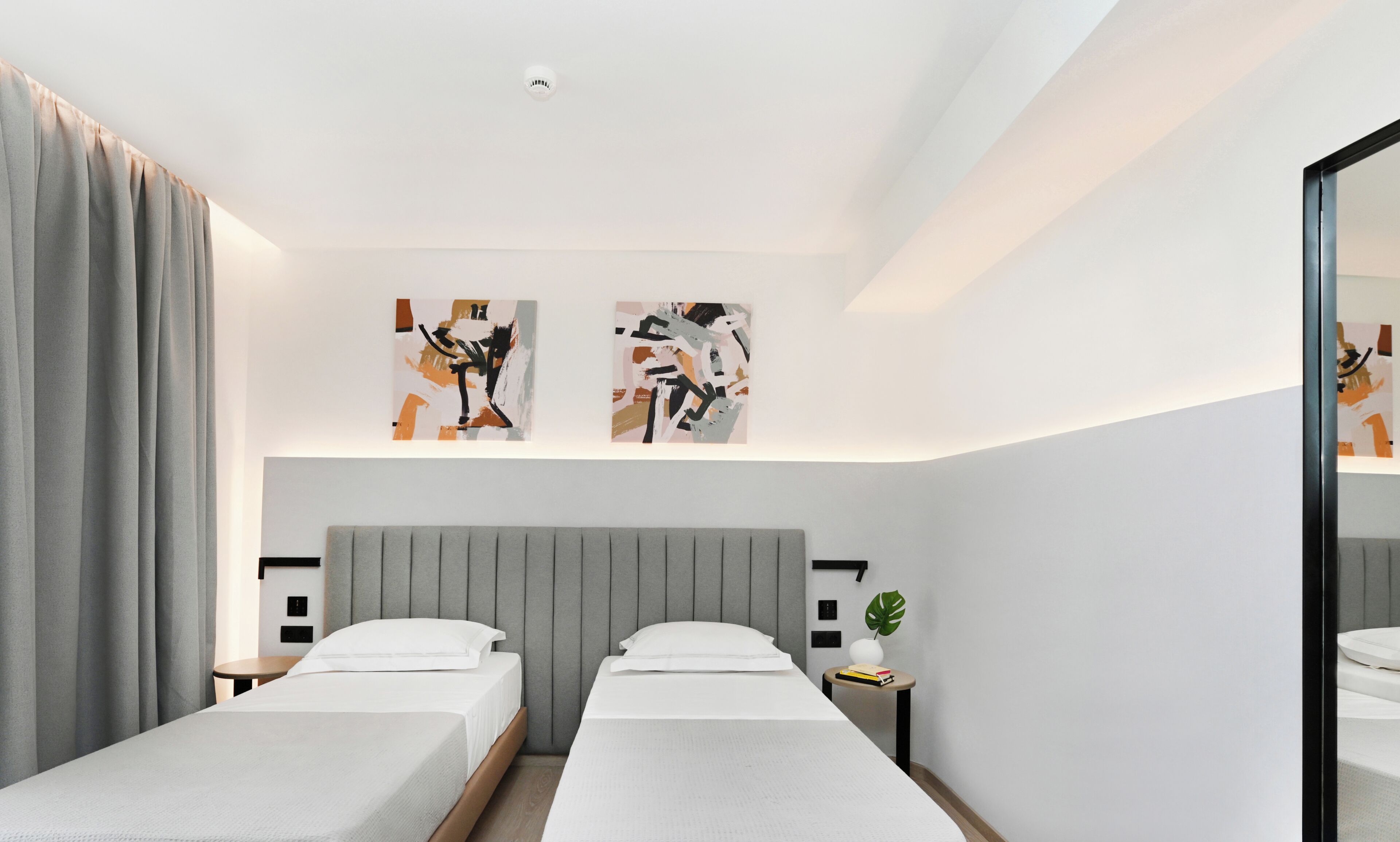 A modern hotel room with two single beds, a grey headboard, a small bedside table with a plant, and abstract art on the wall.