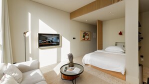 A modern hotel room with a bed, a flat-screen TV mounted on the wall, a round coffee table with a magazine and remote control, and a sofa. - Prime Studio w Burj Views Near Downtown and Canal (Dubai)