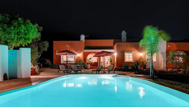 Zona junto a la piscina con sillones de descanso y comedor, rodeada por un edificio de techo azulejado e iluminación cálida.