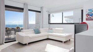 Una sala de estar moderna con un sofá blanco tipo seccional, un televisor de pantalla plana y grandes ventanales con vista al mar.