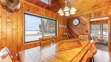 Une salle à manger en bois avec une grande table, des chaises, un lustre, une horloge et une vue sur l’extérieur.