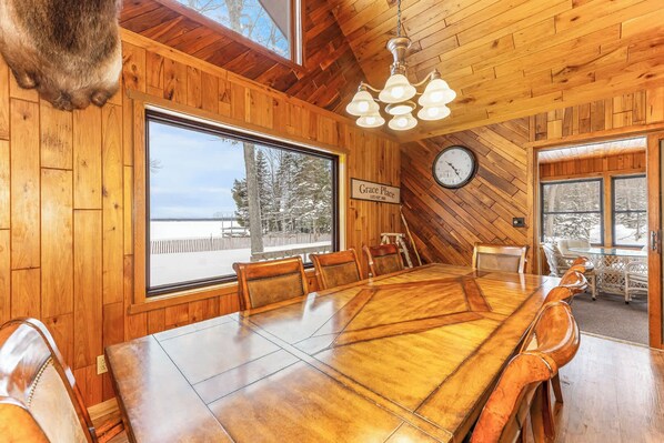 A wooden dining room with a large table, chairs, a chandelier, a clock, and a view of the outdoors.