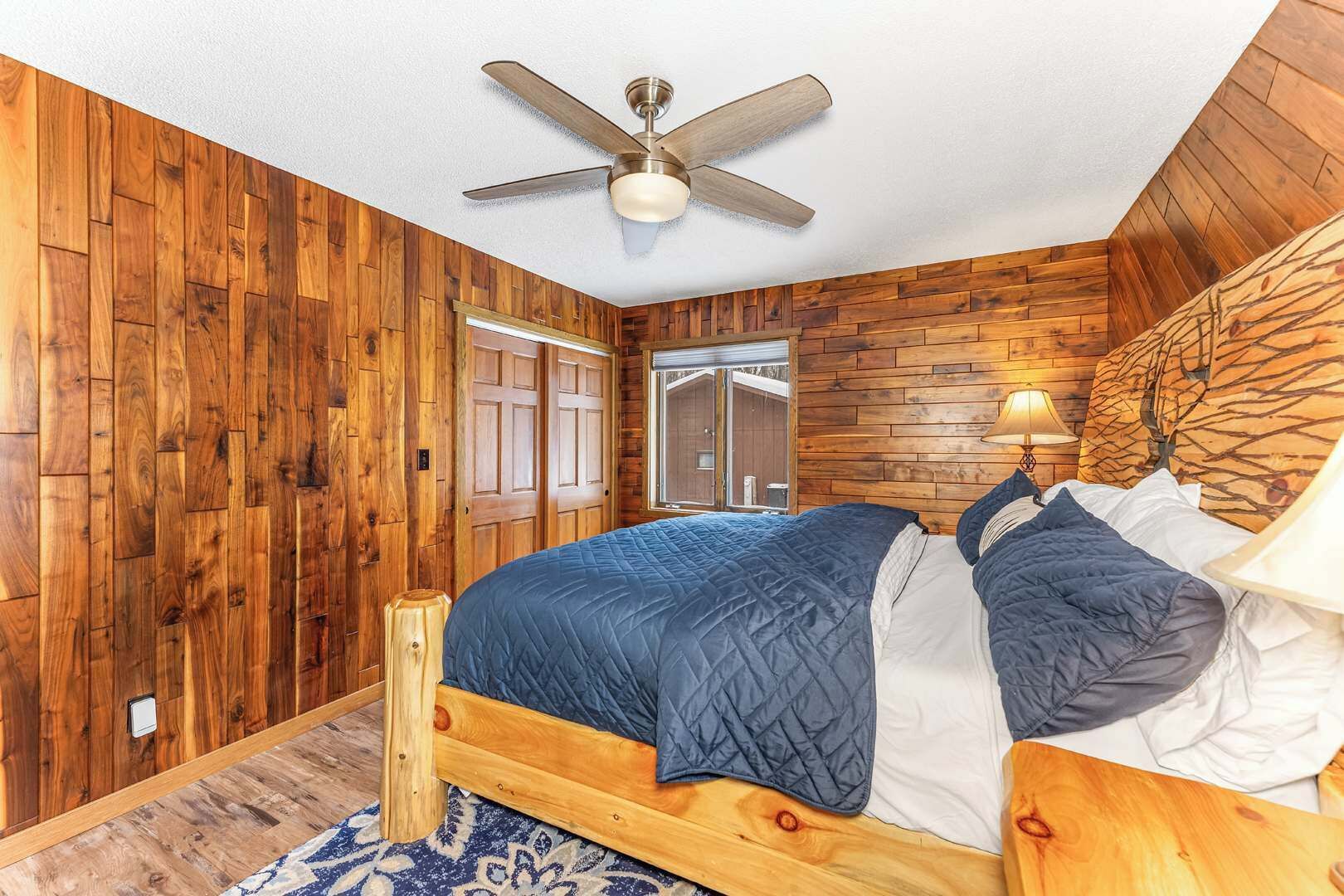 Ein Schlafzimmer mit Holzboden, einem Bett mit blauer Bettwäsche, einem Deckenventilator und einem Fenster mit Blick auf ein Holzgebäude im Freien.
