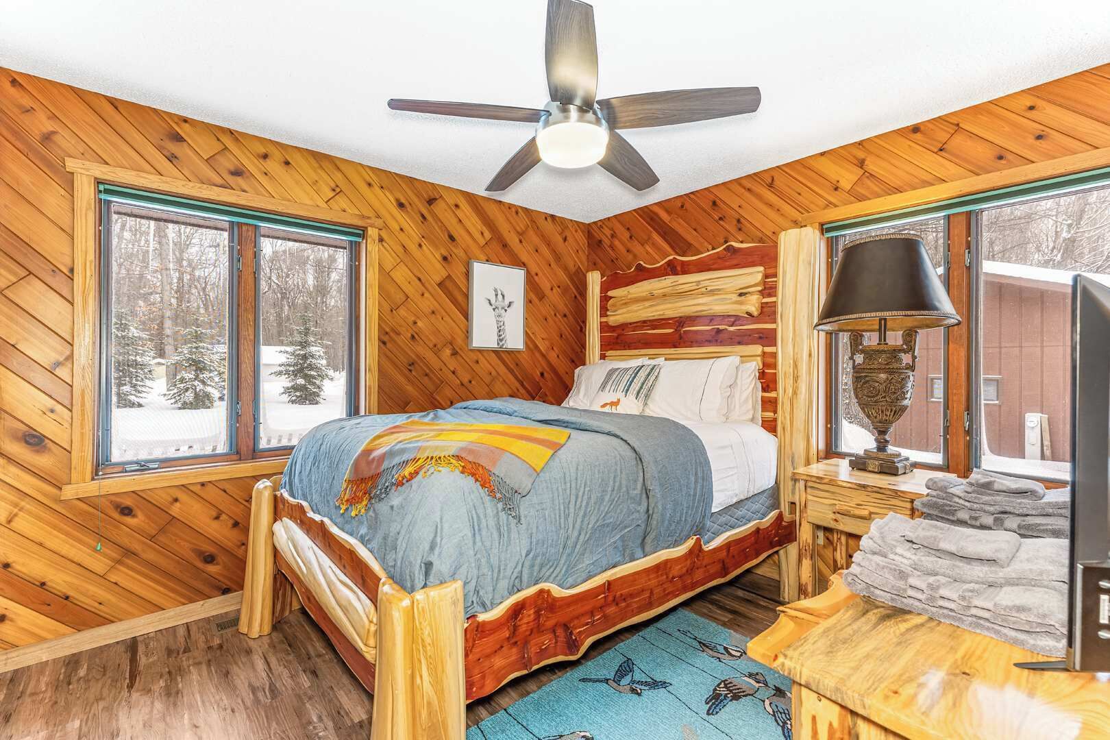 A wooden cabin bedroom with a bed, a ceiling fan, a window with a view of snow-covered trees, and a lamp on a bedside table.