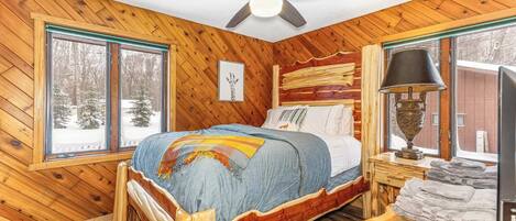 A wooden cabin bedroom with a bed, a ceiling fan, a window with a view of snow-covered trees, and a lamp on a bedside table.