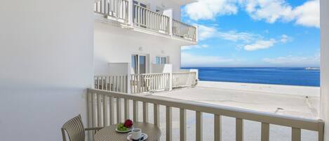 A balcony with a white railing, a round table with a cup and a plate of fruit, and a view of the sea.