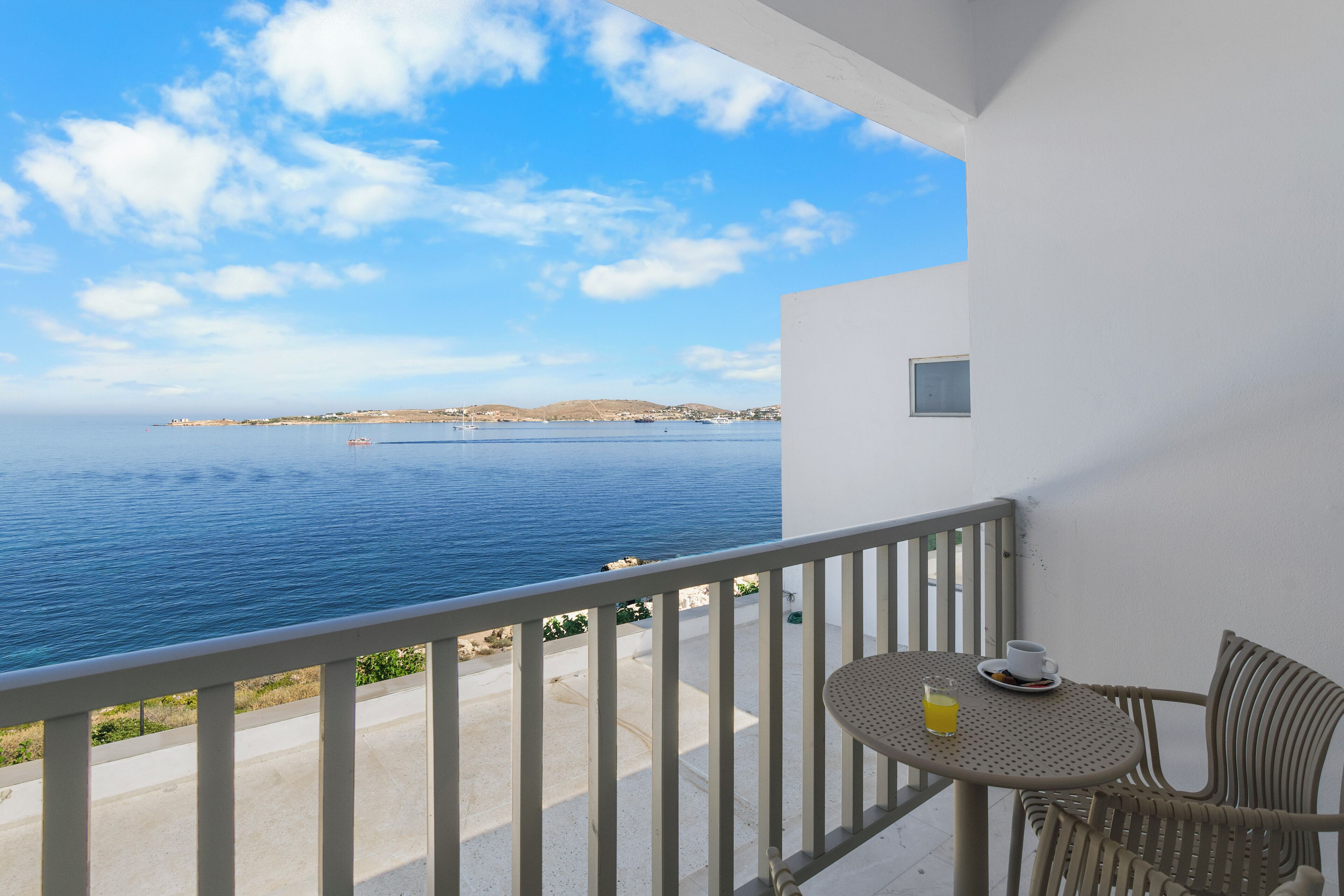 Un balcone con tavolo e sedia, affacciato su una vista costiera con il mare e la terraferma in lontananza.