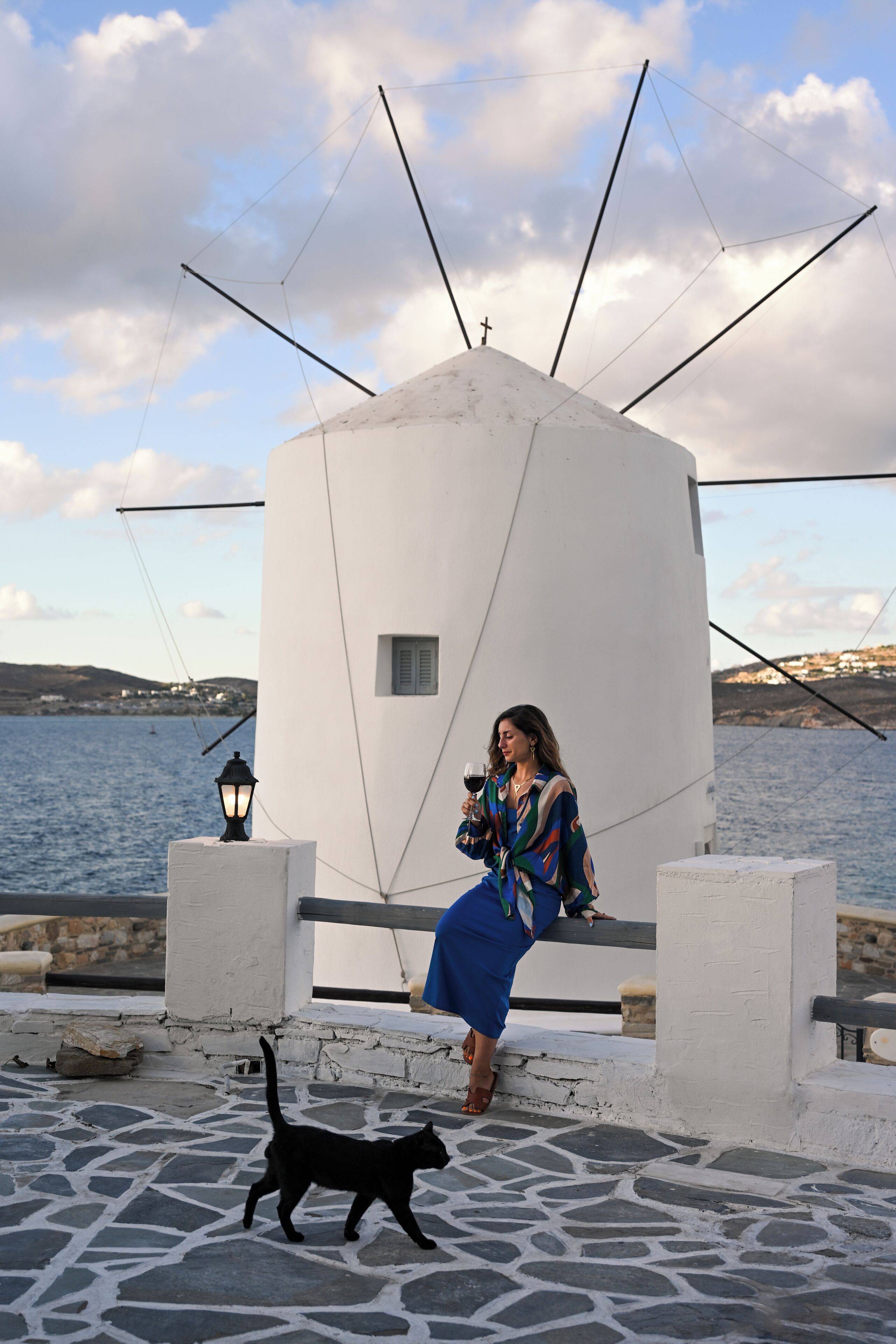 Una mujer sentada en un banco junto a un edificio blanco, con vista al mar.