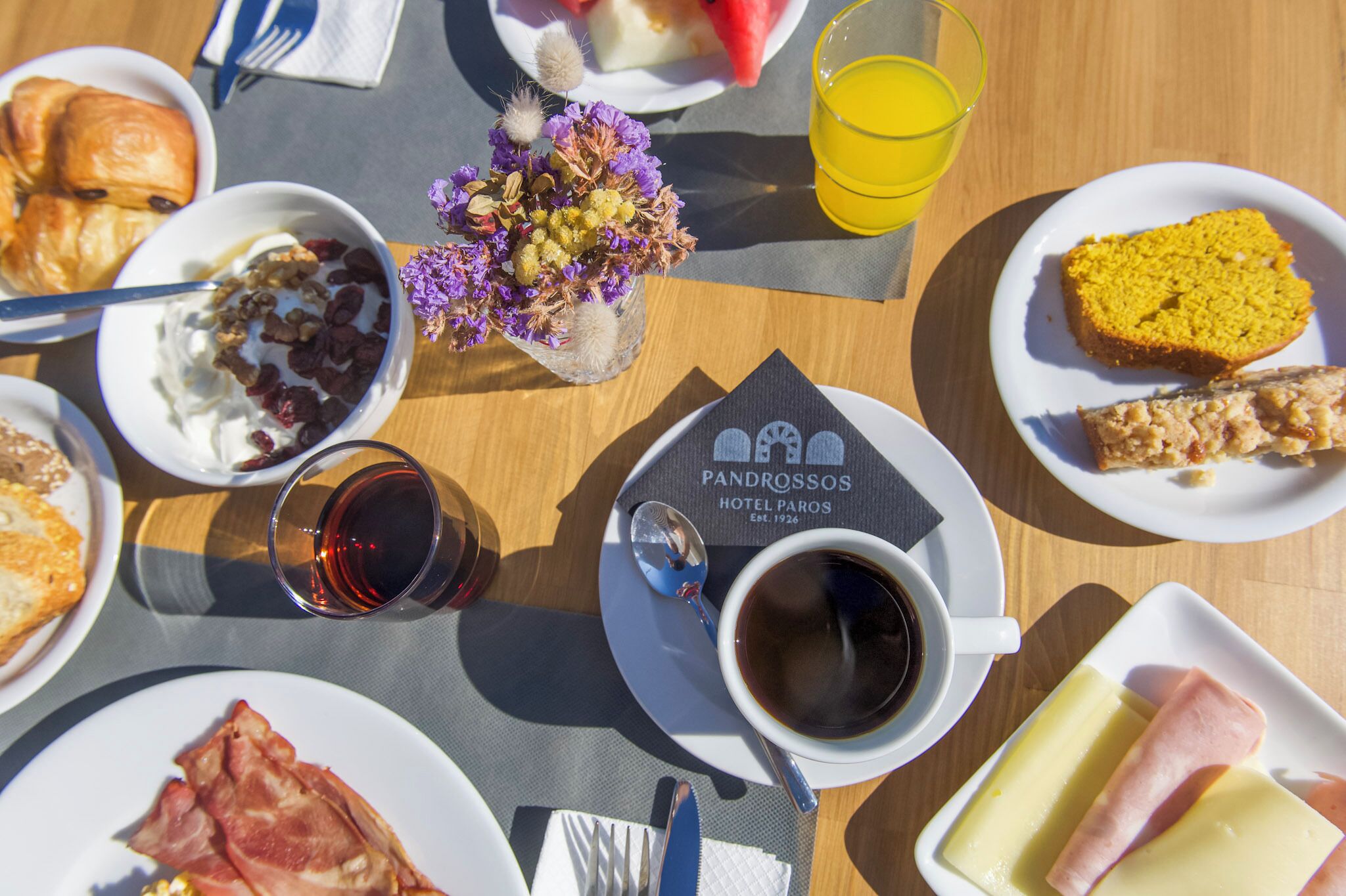 Une table de petit-déjeuner avec des croissants, un yaourt aux fruits rouges, une tranche de pastèque, un verre de jus d’orange, une tasse de café et une assiette de jambon et de fromage.