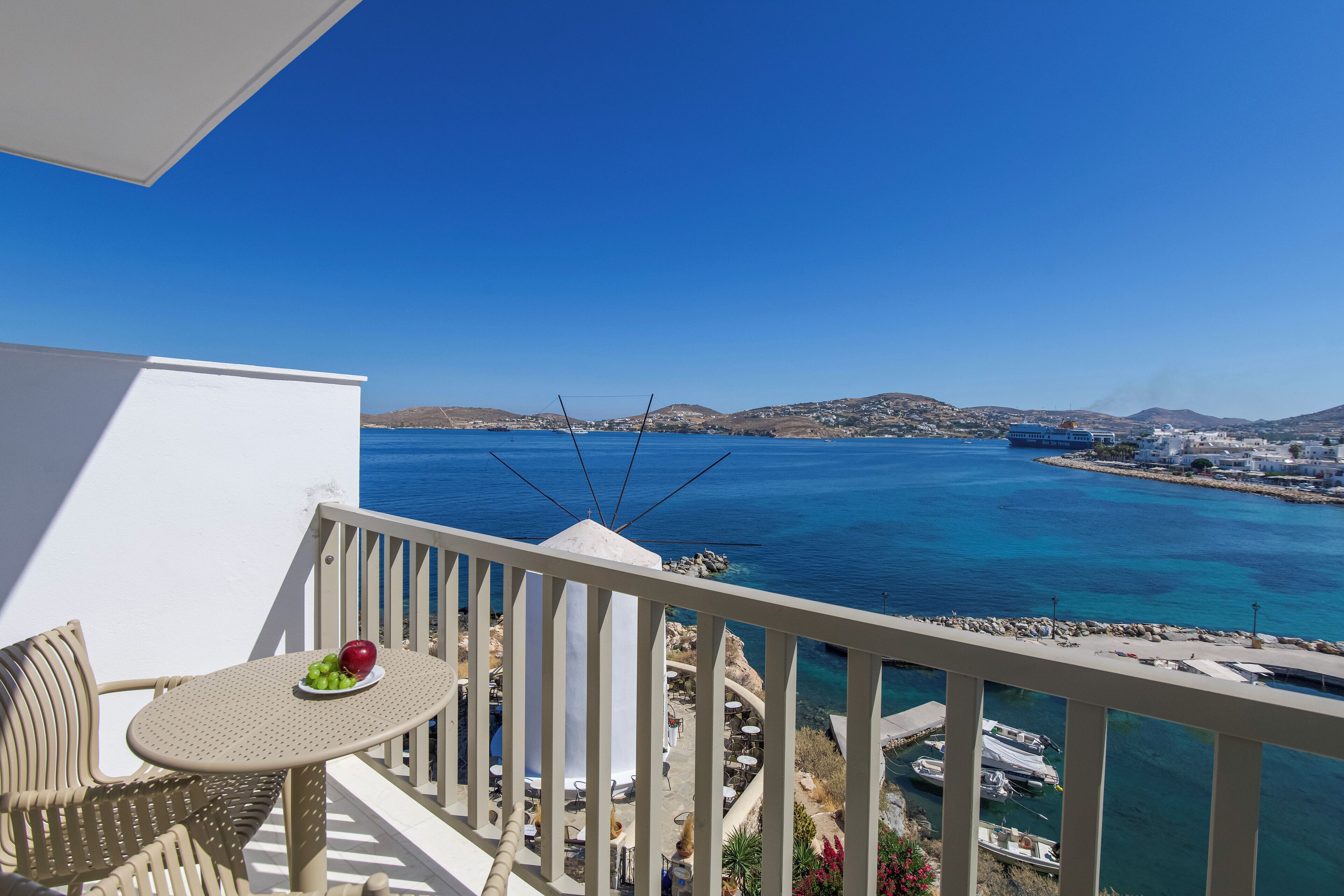 A balcony with a table and chairs, overlooking a coastal area with clear blue water and distant hills.