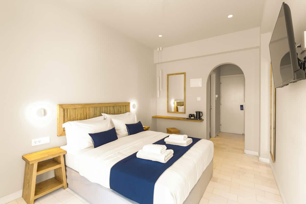 A modern hotel room with a large bed, wooden headboard, white walls, a flat-screen TV, and a small wooden nightstand.