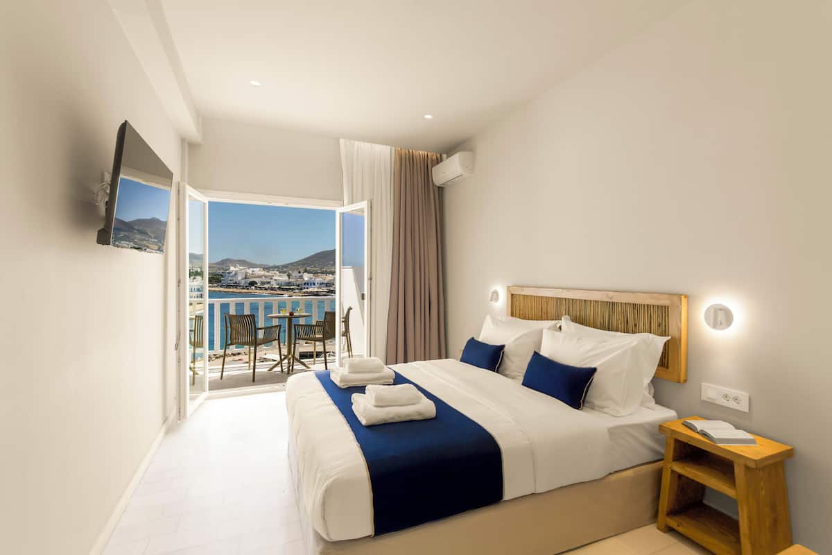 A hotel room with a large bed, a wooden nightstand, a TV mounted on the wall, and a balcony with a view of the city and mountains.