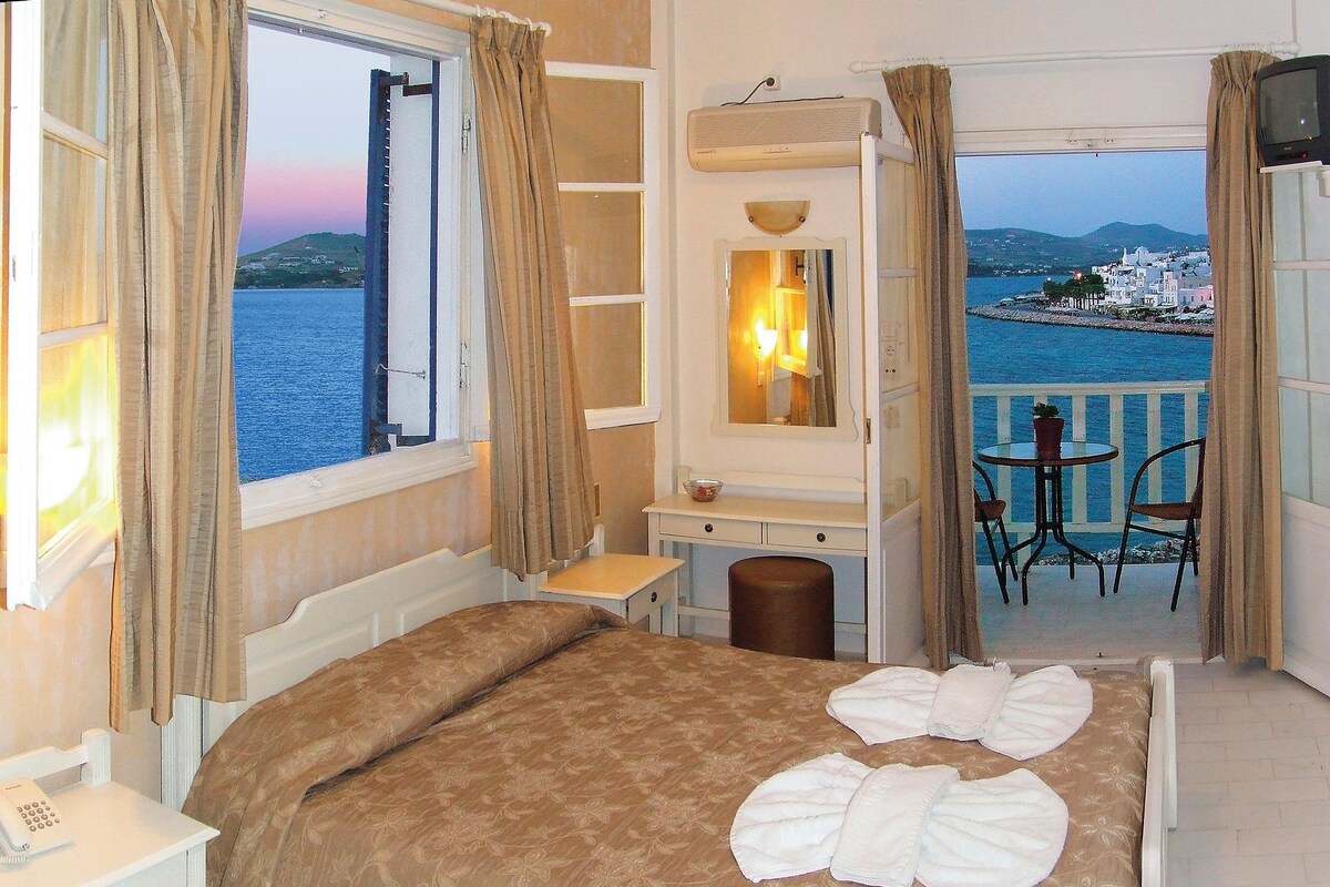 A hotel room with a bed, a desk, a chair, a small table, and a view of the sea and a town.