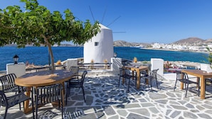 Un área de estar al aire libre con piso de piedra, mesas y sillas, con vista a un paisaje costero que incluye un edificio blanco y el mar.