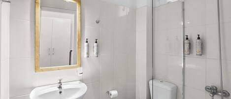 Un baño moderno con lavamanos, espejo y váter blancos, y una zona de ducha con mampara de vidrio.