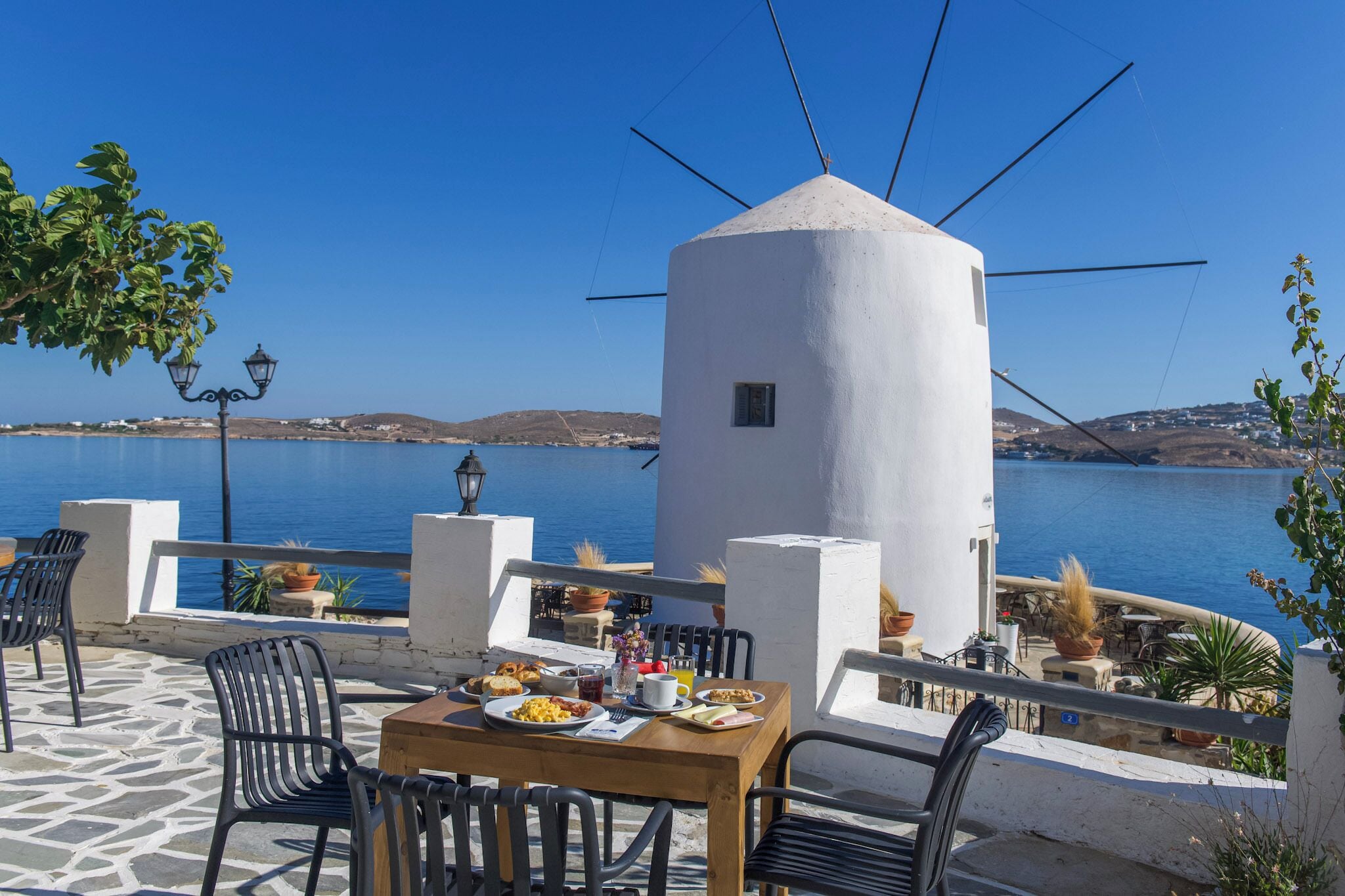 Una terrazza sul mare con un tavolo apparecchiato per mangiare, con vista su un mare tranquillo e un mulino a vento bianco.