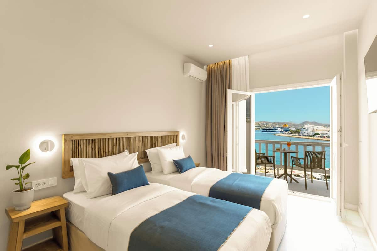 A hotel room with two beds, a balcony with a view of the sea, and a potted plant on the nightstand.