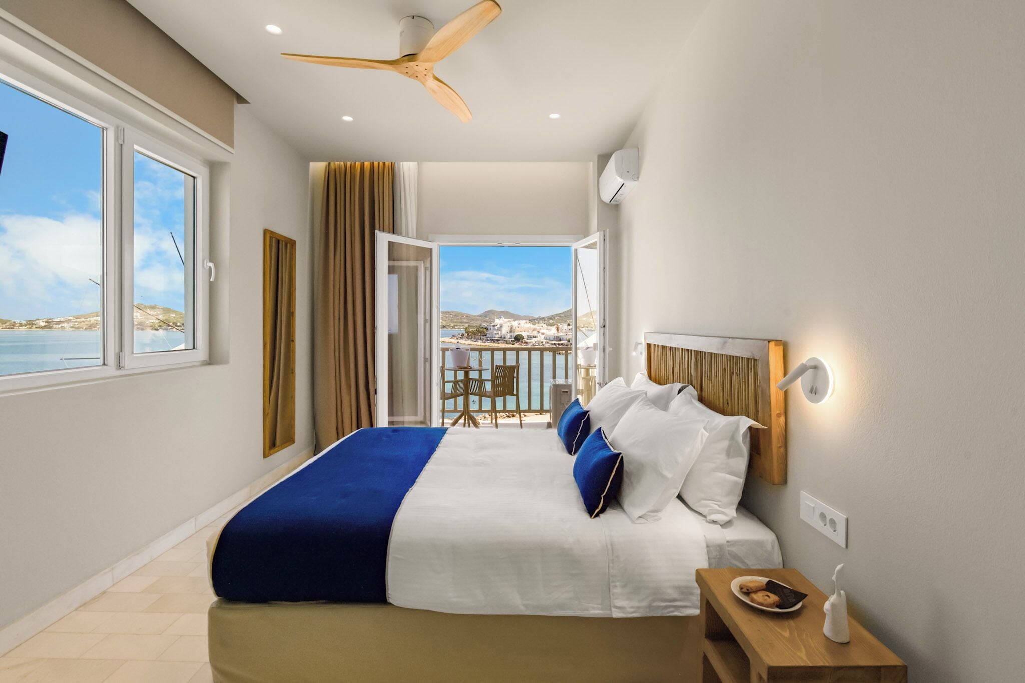 A hotel room with a bed, a ceiling fan, a balcony with a view, and a nightstand with a lamp.