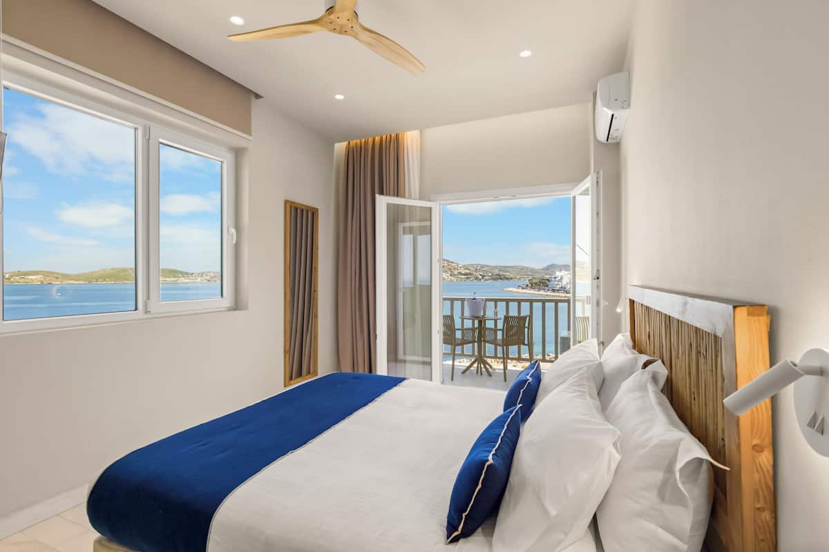 A bedroom with a bed, a ceiling fan, a view of the sea, and a balcony with chairs.