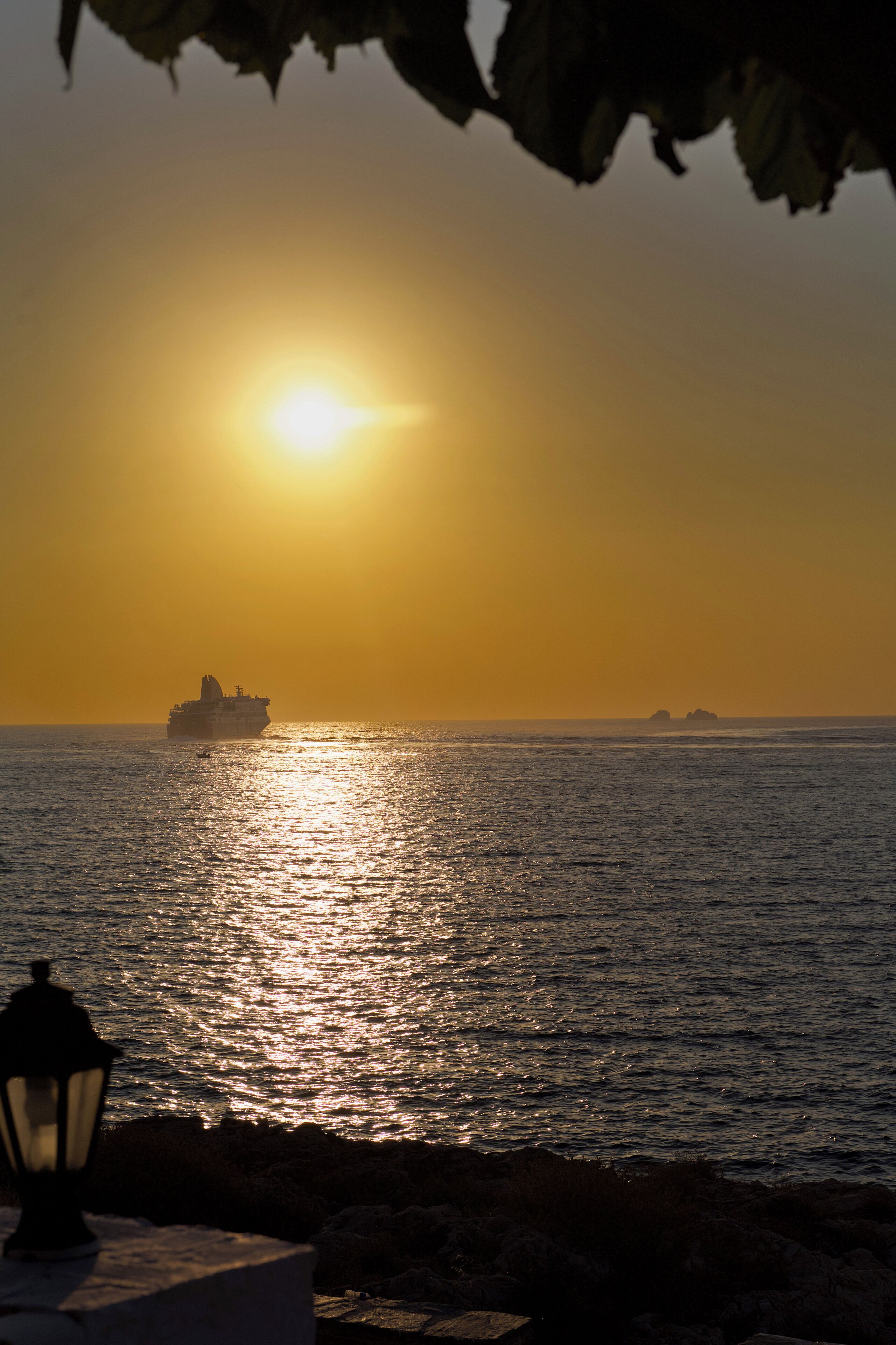 A sunset over the ocean with a ship in the distance and a lamp post in the foreground.