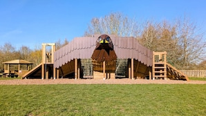 A wooden play structure with a bird-like roof, a slide, and a climbing ladder. - Oak Lodge (Swadlincote)