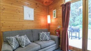 A cozy cabin room with a wooden interior, a gray sofa with patterned cushions, a floor lamp, a fire extinguisher, and a view of the outdoors through a glass door. - Oak Lodge (Swadlincote)