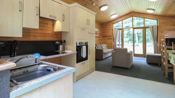 A modern kitchen with granite countertops, stainless steel appliances, and wooden cabinetry. There is a dining area with a table and chairs, and a large window providing a view of the outdoors. - Rowan Lodge (Swadlincote)