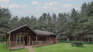 A wooden cabin with a porch, a picnic table, and a grassy area surrounded by trees. - Rowan Lodge (Swadlincote)