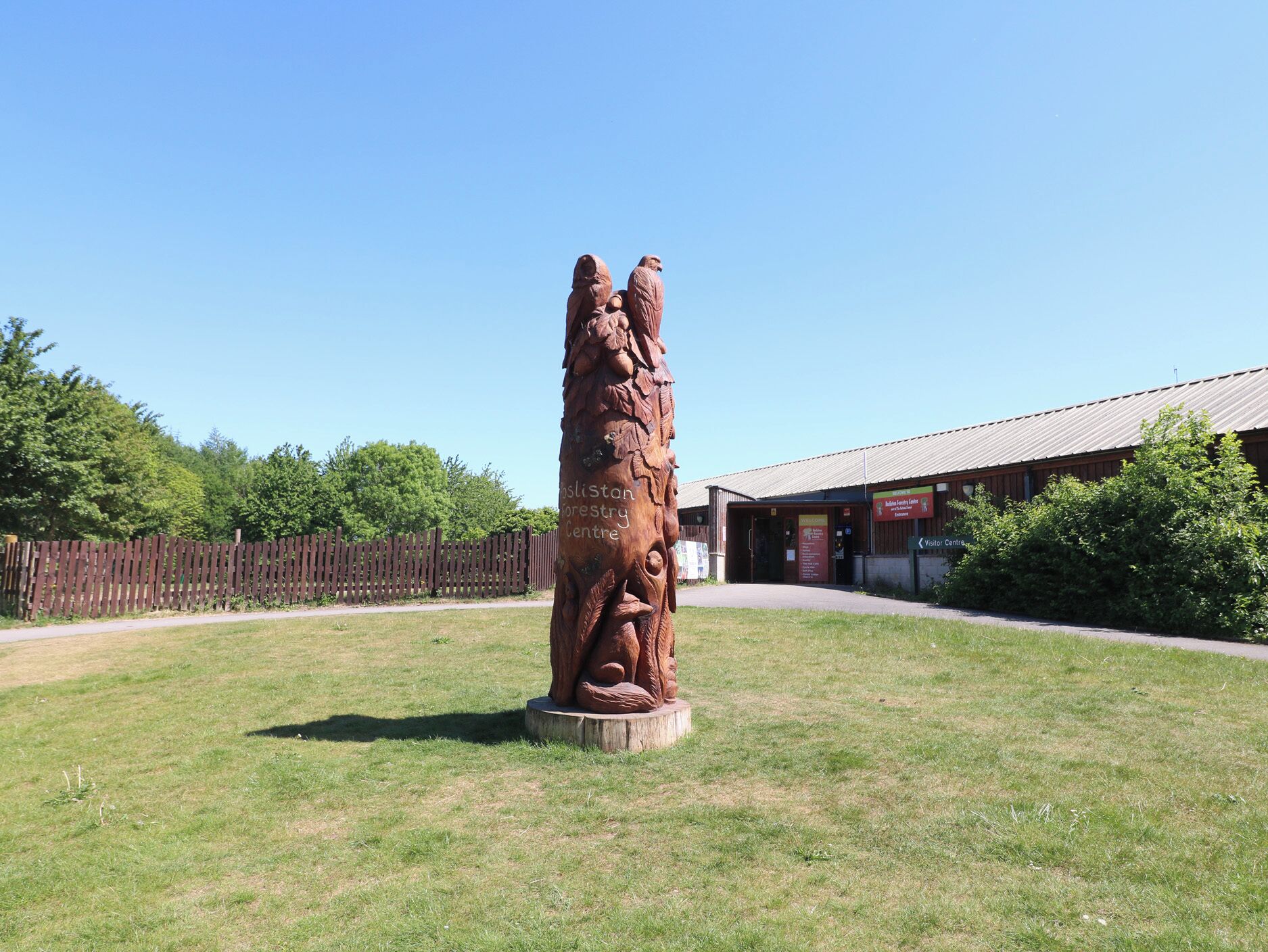 Une sculpture en bois, située dans un espace verdoyant, avec un bâtiment en arrière-plan.