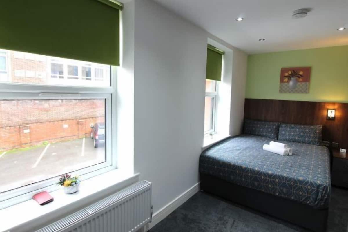 A bedroom with a bed, a radiator, a window with a view of a brick wall and a car, and a wall-mounted light fixture.