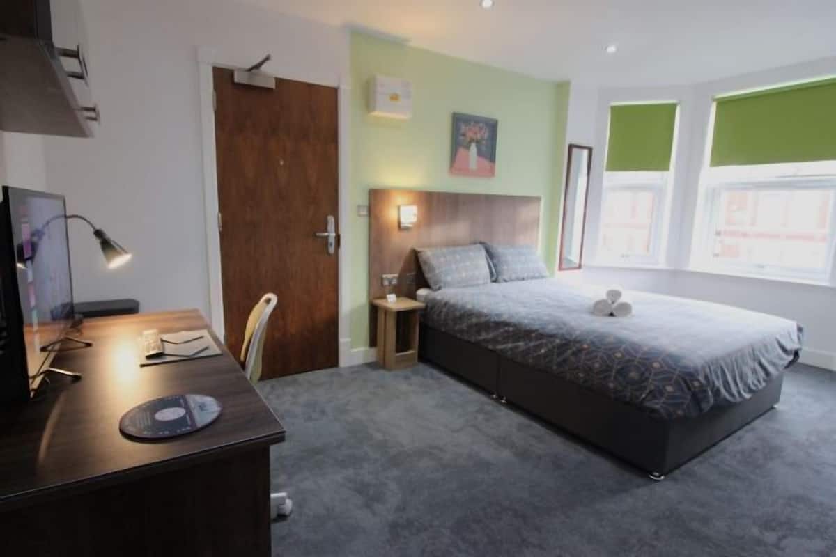 A bedroom with a bed, bedside table, lamp, desk, chair, and a window with blinds.