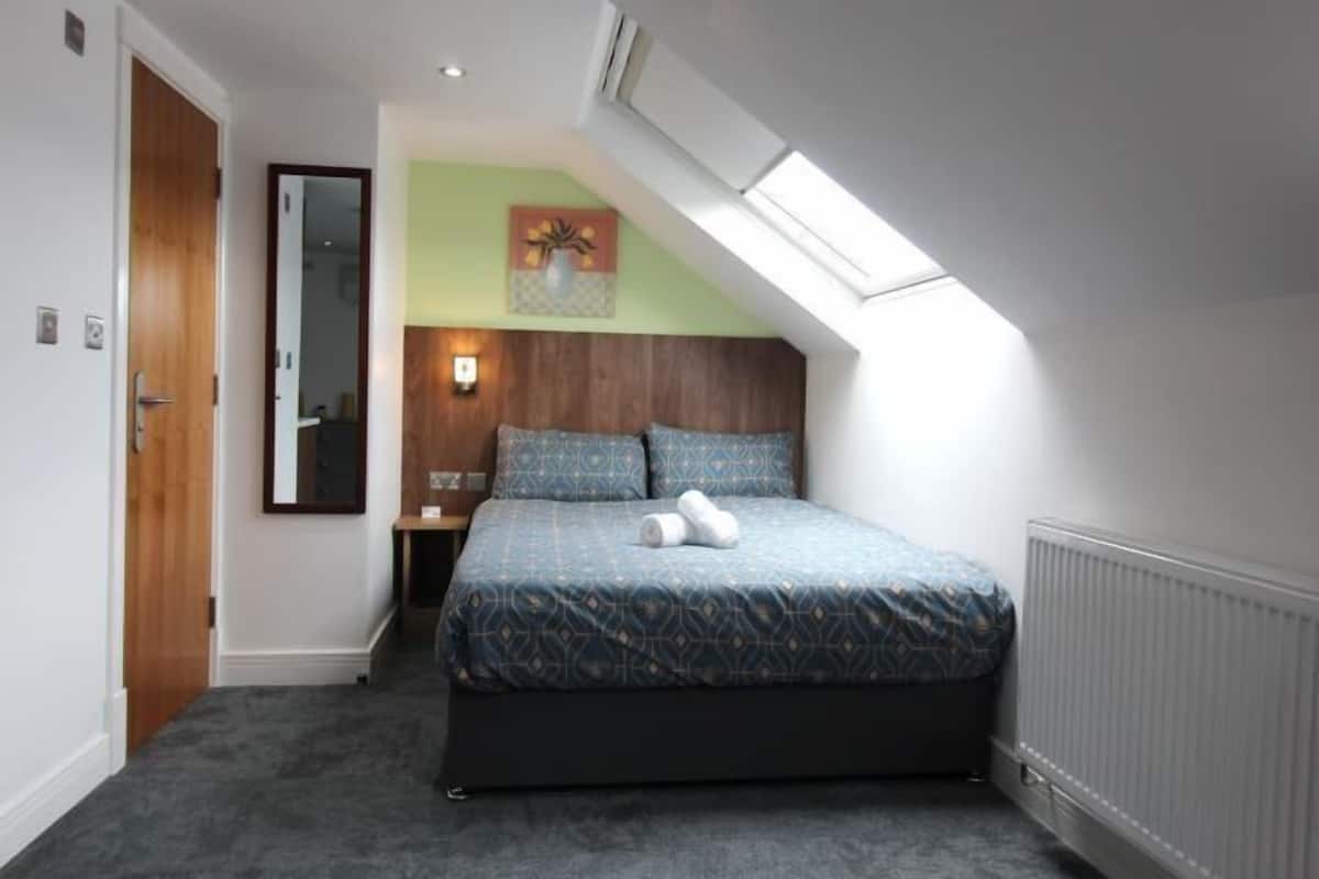 A bedroom with a sloped ceiling, a bed with patterned bedding, a nightstand, a wall-mounted lamp, and a painting.