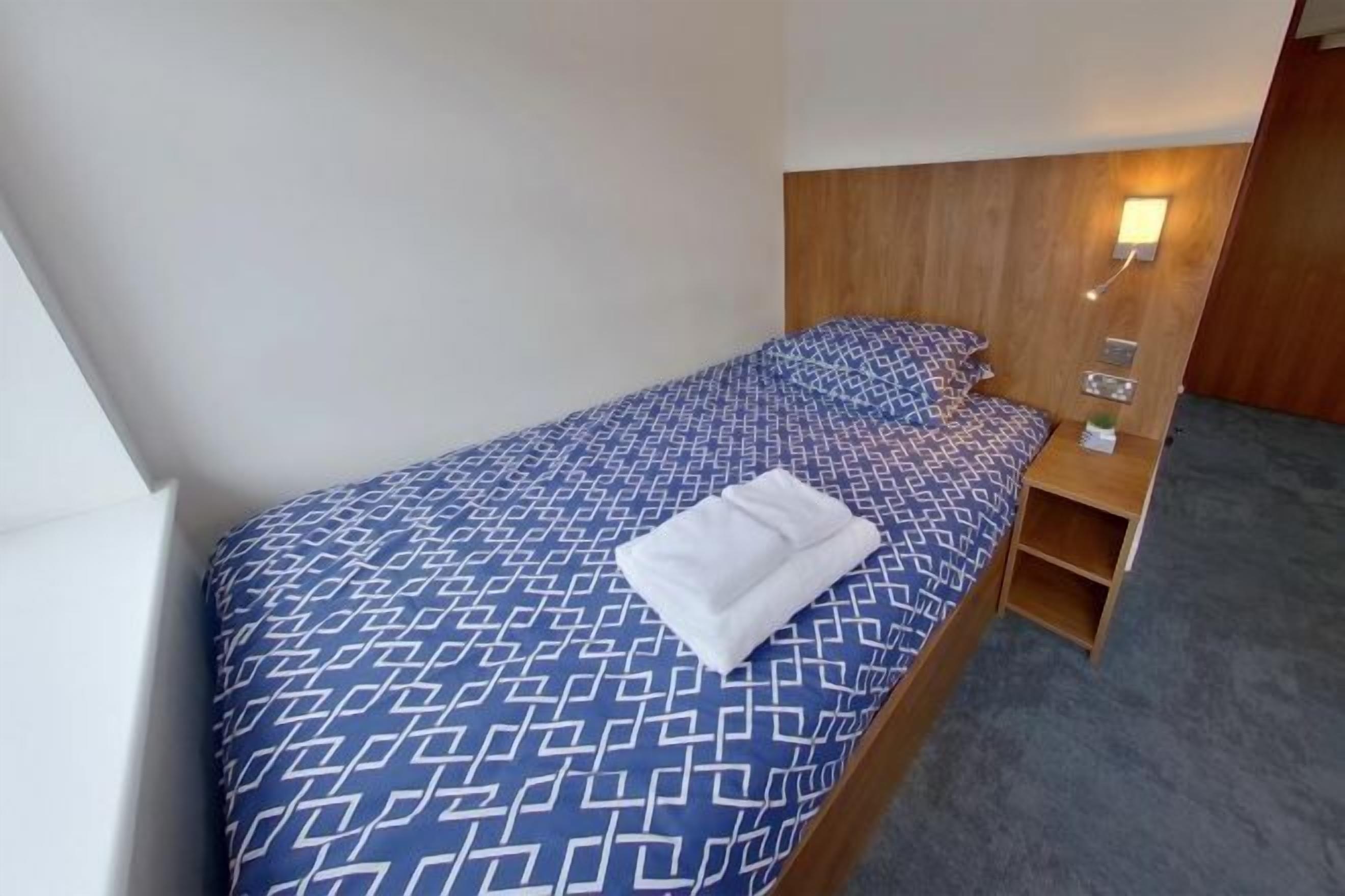 Ein Einzelbett mit geometrischem Muster auf der Bettdecke, ein hölzernes Kopfstück, ein Nachttisch mit Lampe und eine kleine Ablagefläche.