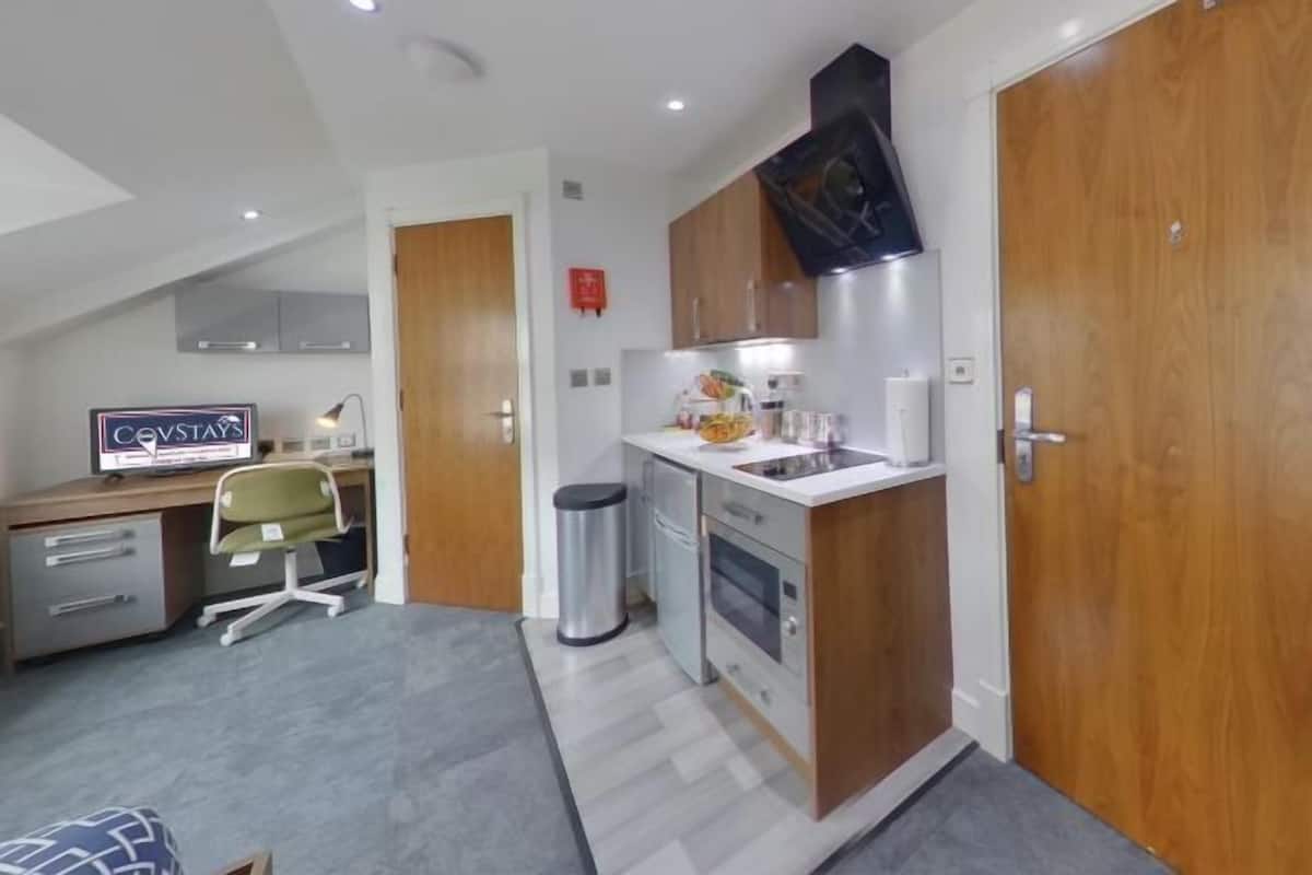 A compact kitchen with a microwave, oven, and sink, a small dining area with a chair, and a desk with a computer and a sign.
