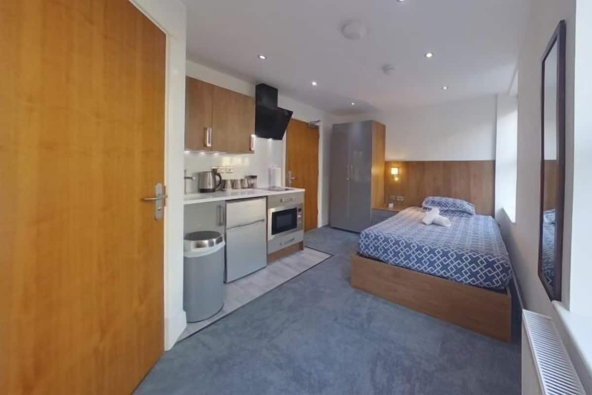 A modern hotel room with a kitchenette, a bed with patterned bedding, and a large window.