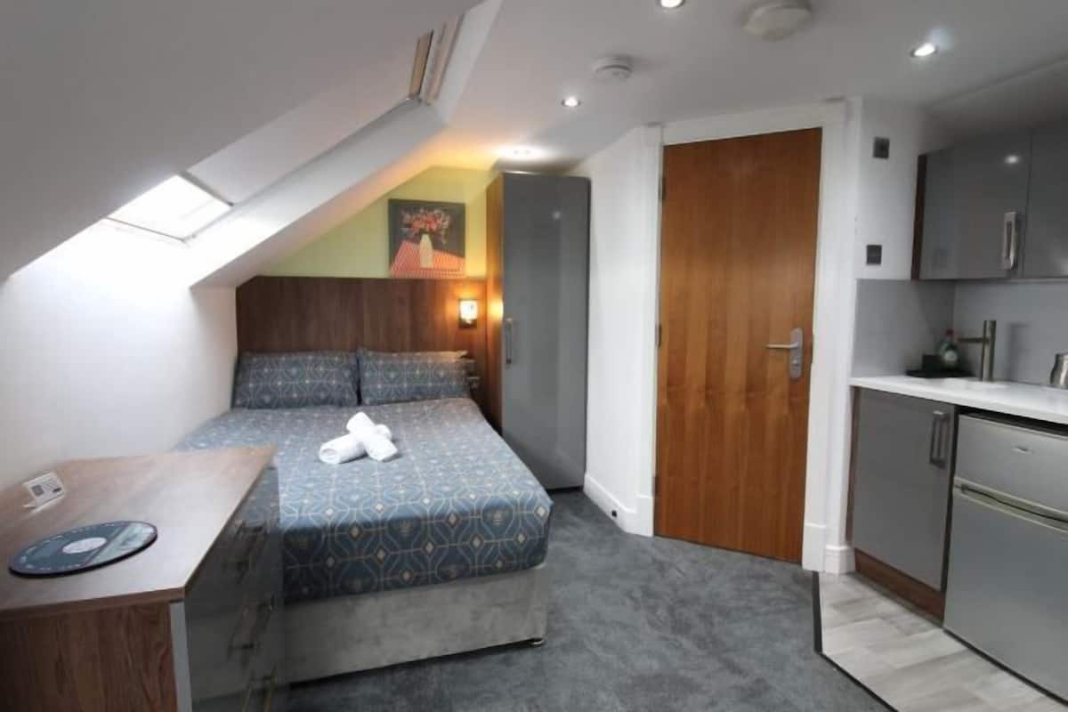 A compact room with a bed, a small desk, a kitchenette, and a door leading to another room.
