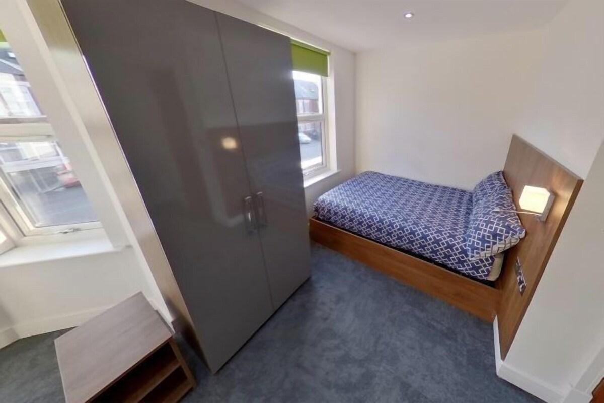 A small bedroom with a bed, a wardrobe, a window, and a small wooden shelf.