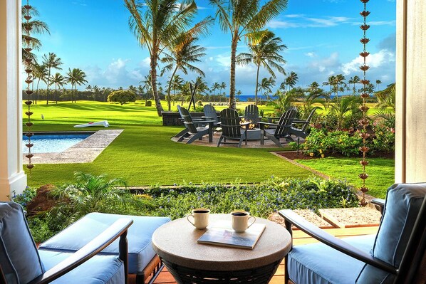 A view from a balcony overlooking a well-manicured lawn with a pool, outdoor seating area, and a golf course in the background. - Perfection in Poipu: Brand new luxury home, ocean views, pool+spa (Koloa)