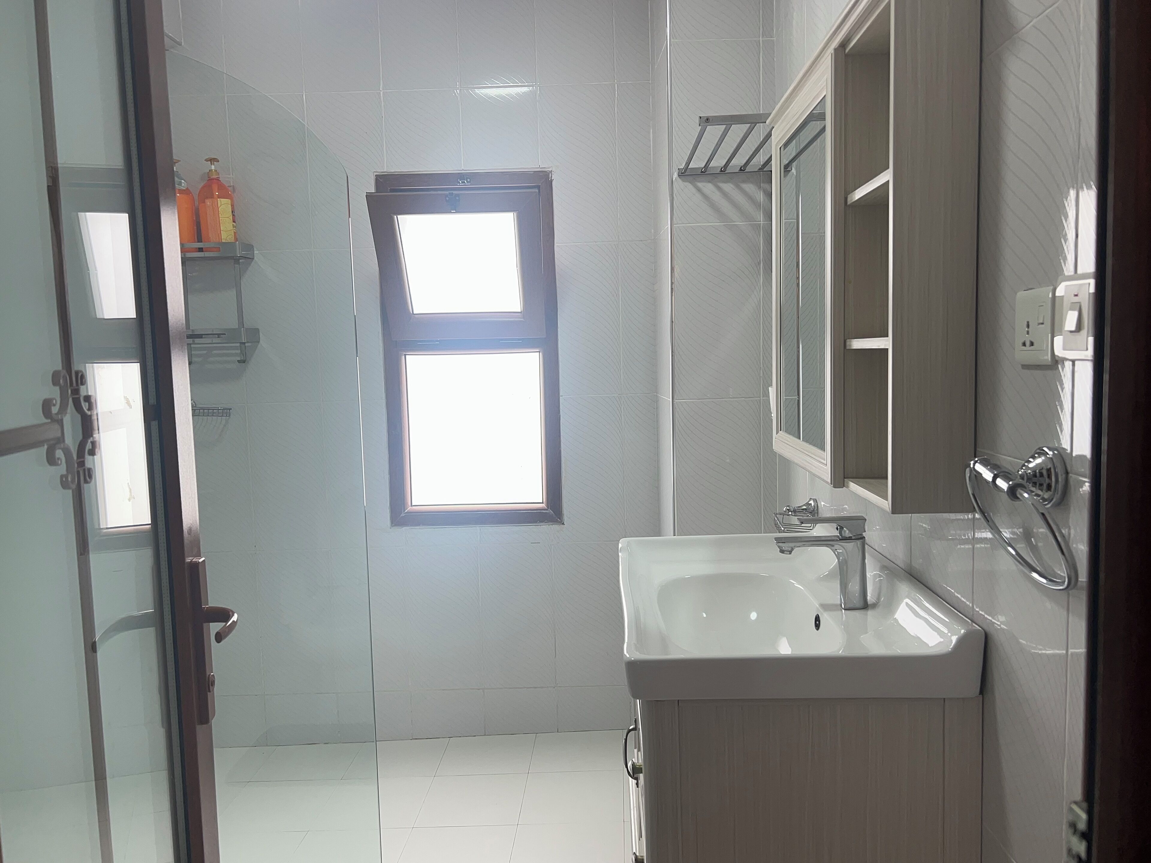 A bathroom with a white sink, a mirror, and a window. There are toiletries on a shelf.