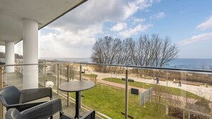 A balcony with a glass railing, a round table, and two chairs. The view includes a grassy area, a walking path, and a body of water. - Apartamenty Sun & Snow Marine (Kolobrzeg)