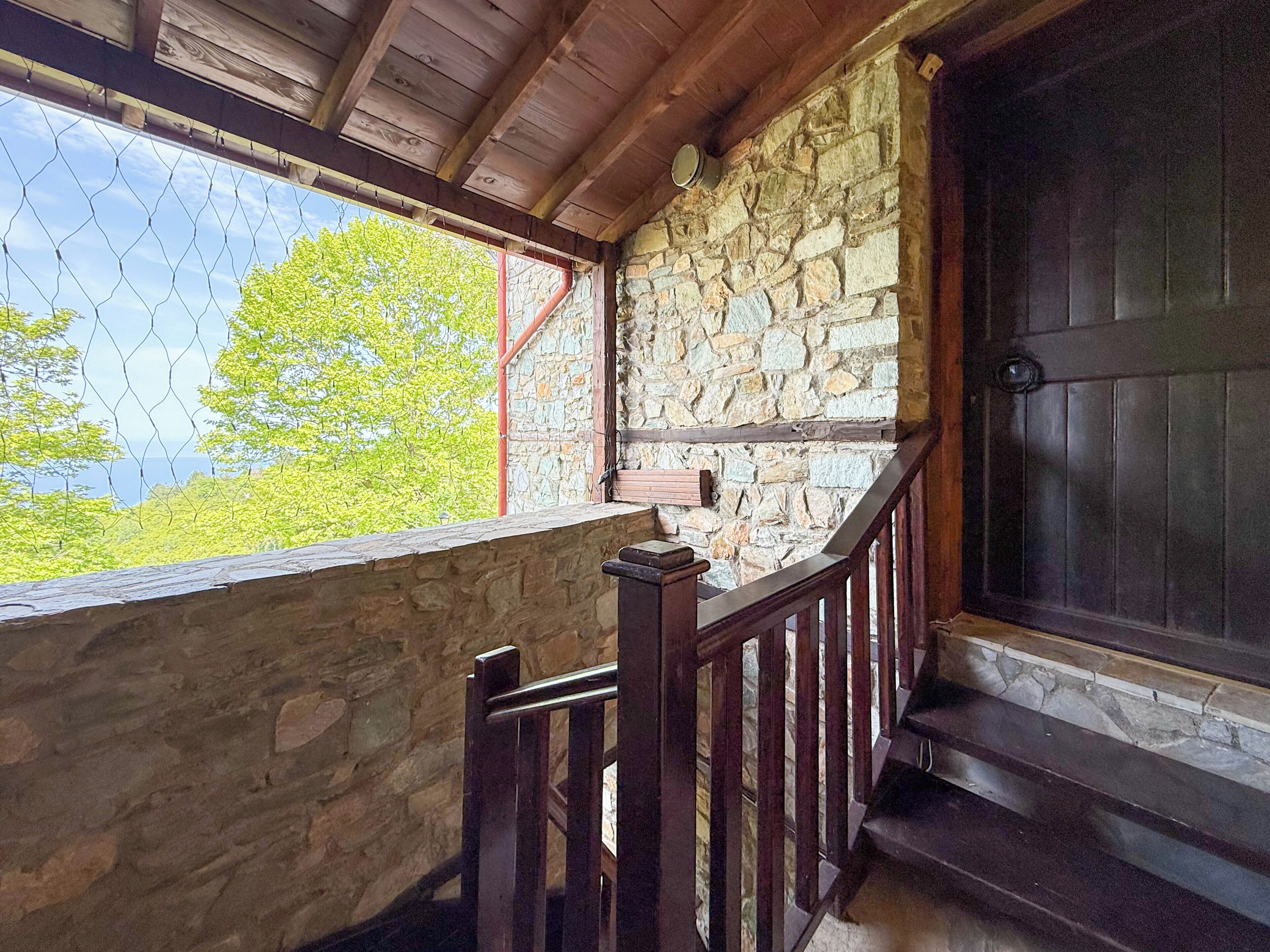 A stone-walled balcony with a wooden railing, a view of green trees, and a dark wooden door.