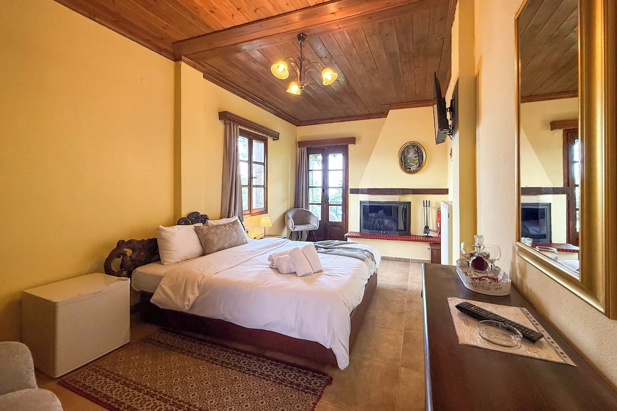 Une chambre avec un plafond en bois, un lit recouvert de draps blancs, une petite table avec une télécommande et une cheminée.