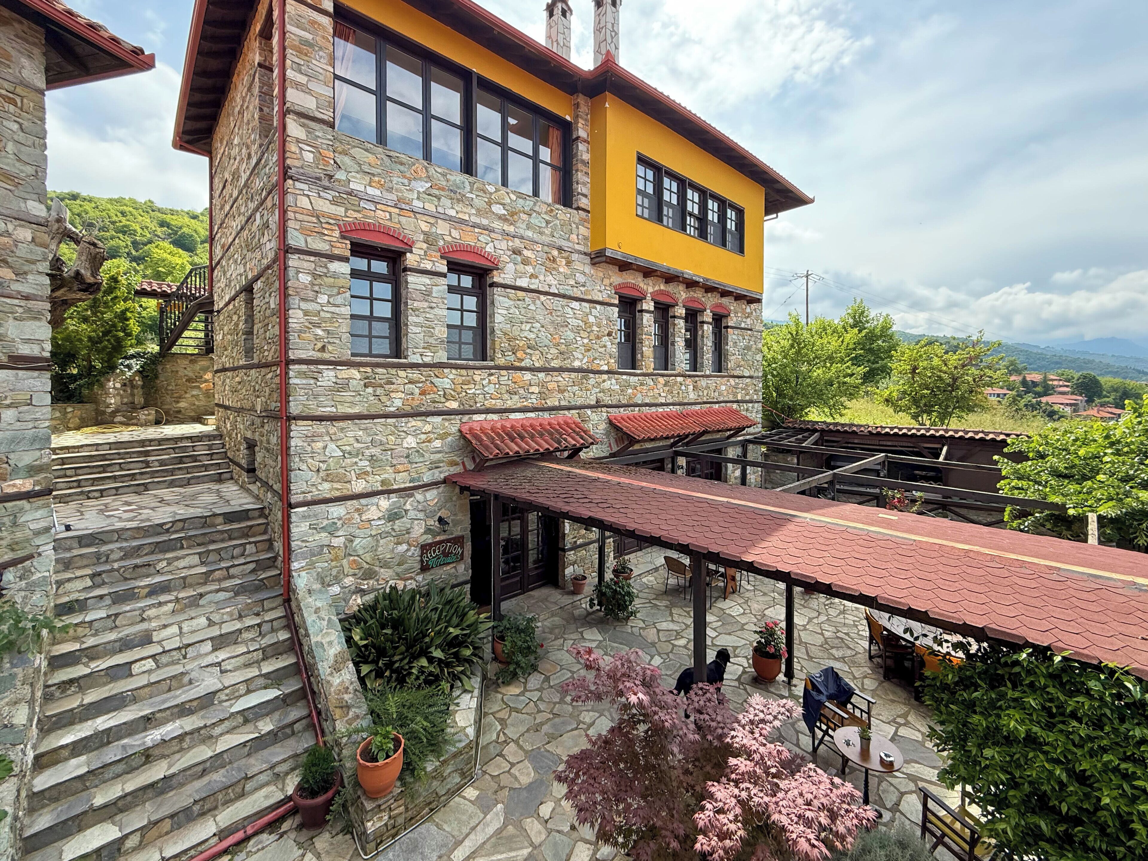 Un edificio de piedra con una sección superior amarilla, escalones de piedra que conducen a un patio cubierto con mobiliario exterior y macetas con plantas.