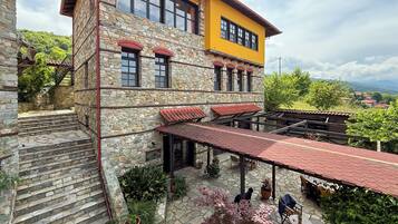 Un edificio de piedra con una sección superior amarilla, escalones de piedra que conducen a un patio cubierto con mobiliario exterior y macetas con plantas.