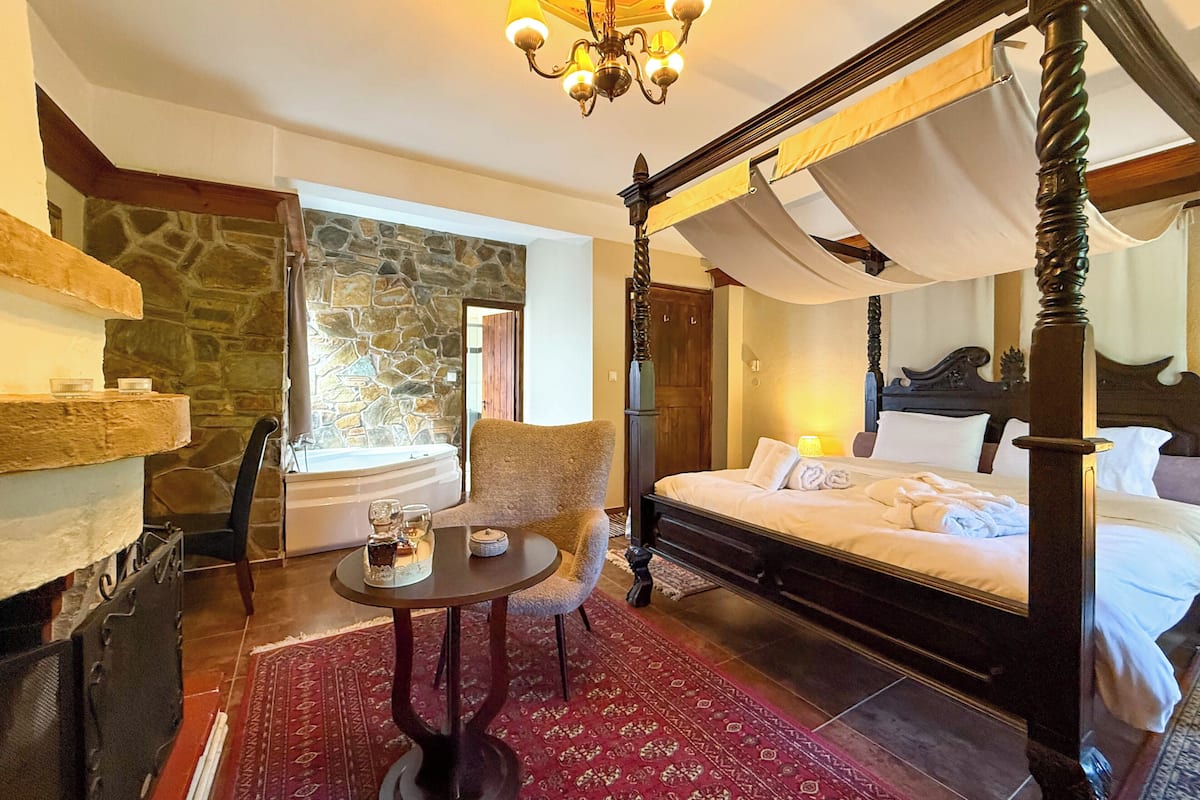 Un lit à baldaquin, un mur de pierre, une chaise en bois, une petite table ronde avec une bougie, et une vue sur un bâtiment en pierre à l’extérieur.