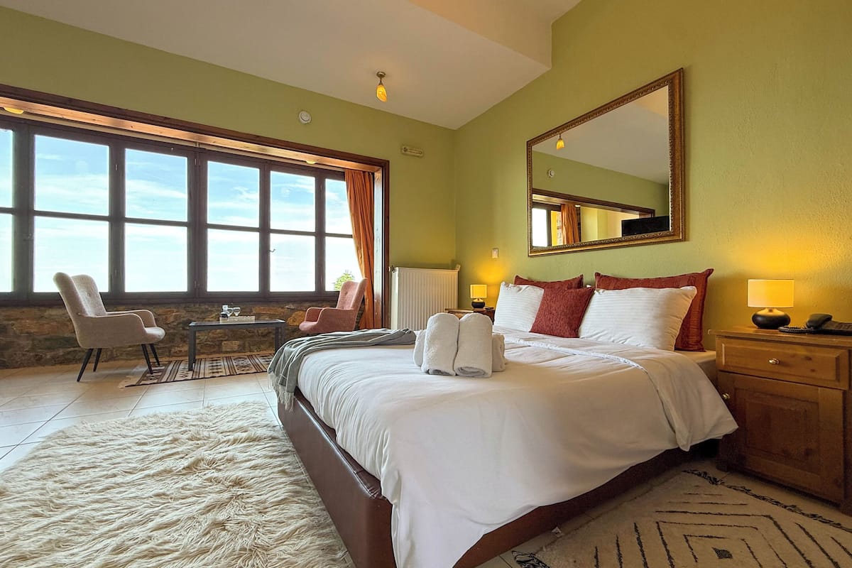 Une chambre d’hôtel avec un grand lit, un coin salon avec un fauteuil et une table, une grande fenêtre avec des rideaux et un miroir au mur.