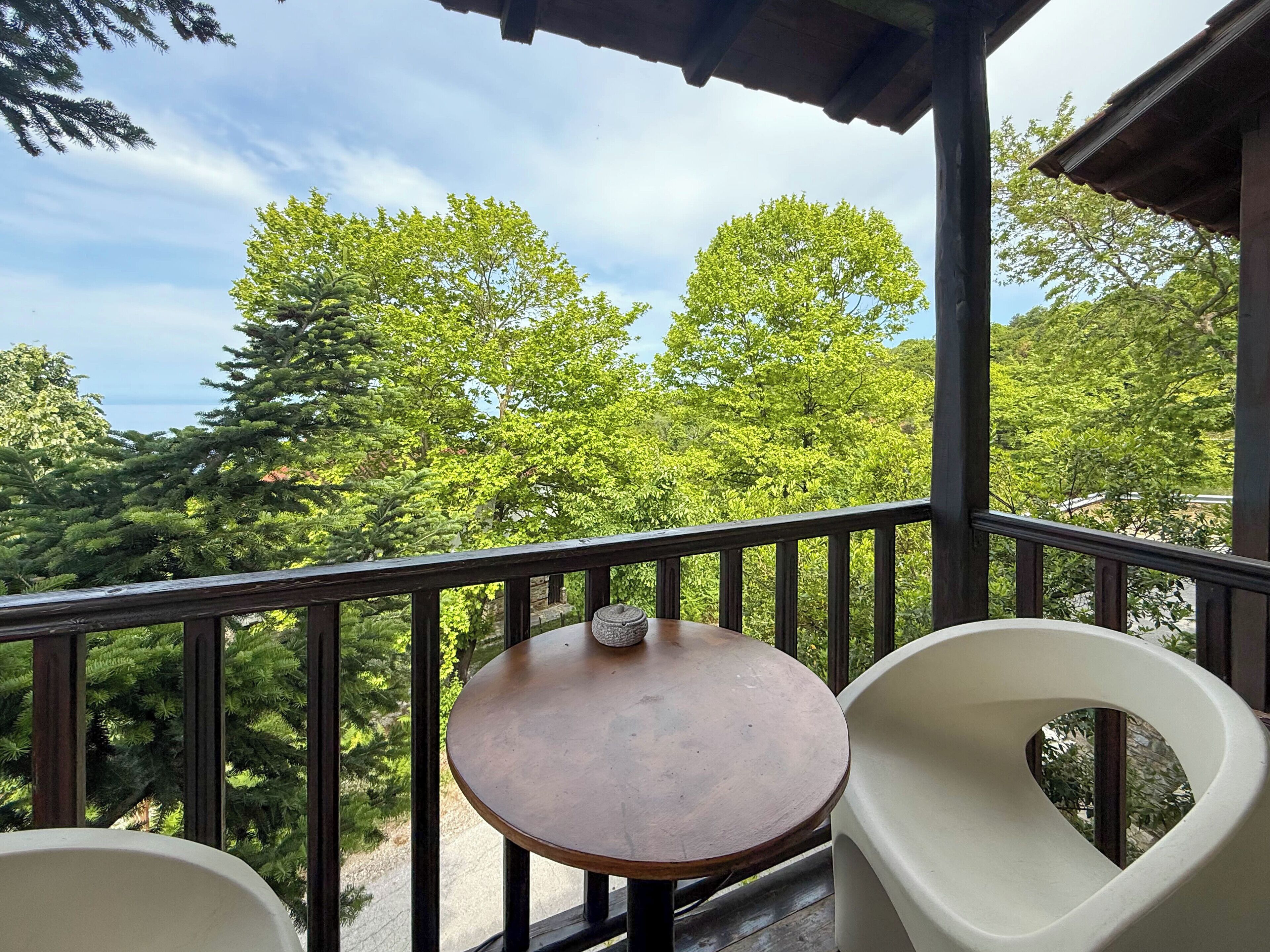 Ein Balkon mit einem runden Holztisch und zwei weißen Stühlen, mit Blick auf eine üppige grüne Landschaft mit Bäumen und blauem Himmel.